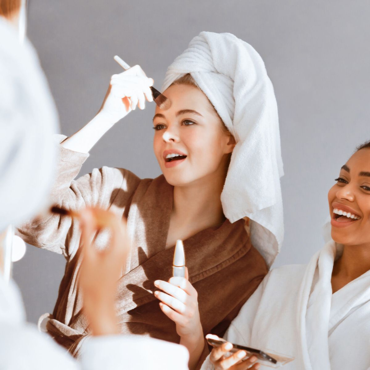 Cheerful multiethnic girls in bathrobes standing in front of big mirror, applying make up foundation with brushes
